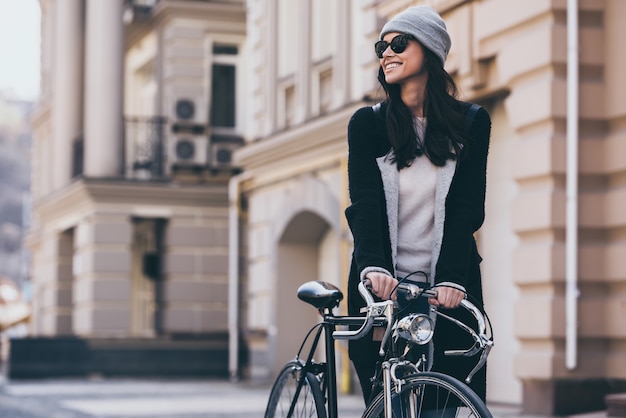 Profiter de cette belle journée. Belle jeune femme à lunettes de soleil roulant son vélo et détournant les yeux avec le sourire en marchant à l'extérieur