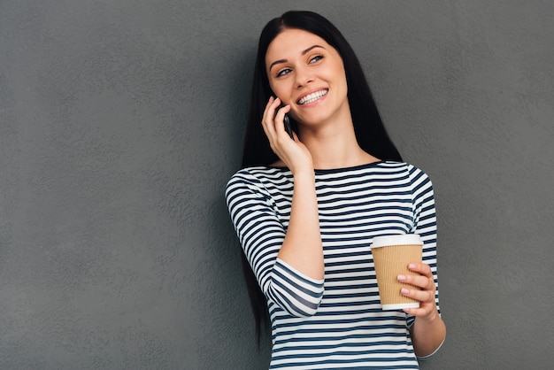 Profiter d'une bonne conversation et d'une boisson chaude. Jolie jeune femme tenant une tasse de café et souriant tout en parlant au téléphone portable et debout