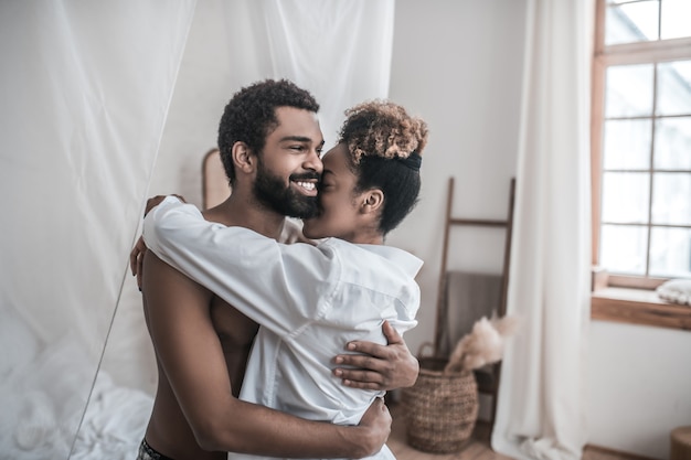 Profiter du moment. Jeune homme américain barbu adulte embrassant tendrement sa femme bien-aimée debout dans une pièce lumineuse