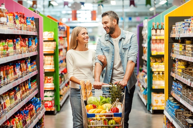 Profiter du shopping ensemble. Heureux jeune couple se liant les uns aux autres et souriant en marchant en marchant dans un magasin d'alimentation avec un panier