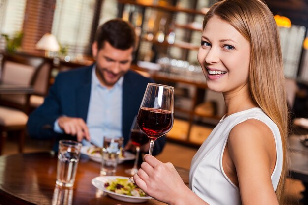 Profiter du temps ensemble. Belle jeune femme tenant un verre de vin rouge et souriante assise au restaurant avec son petit ami assis en arrière-plan