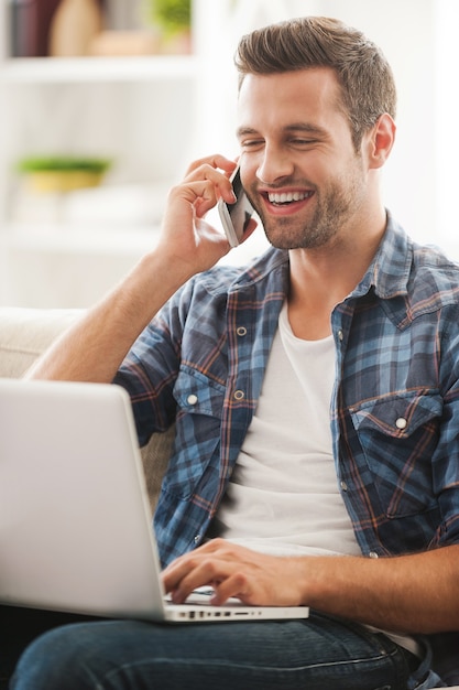 Profiter du temps en ligne. Heureux jeune homme travaillant sur un ordinateur portable et parlant au téléphone assis sur un canapé