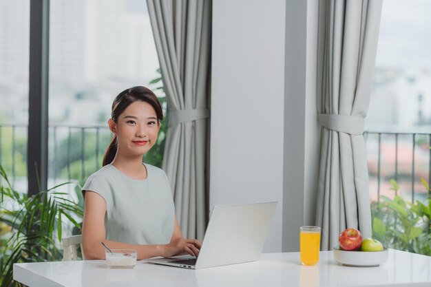 Profiter du temps à la maison Belle jeune femme souriante travaillant sur un ordinateur portable et buvant du jus d'orange