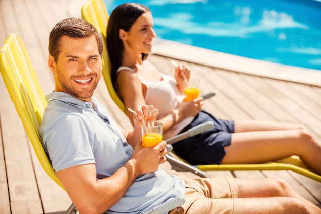 Profiter de leur temps libre ensemble. Vue latérale d'un beau jeune couple assis sur les chaises longues au bord de la piscine et buvant des cocktails pendant que l'homme regarde la caméra et sourit