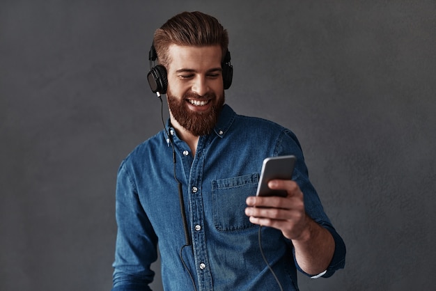 Profiter de la musique préférée. Beau jeune homme tenant un téléphone intelligent et le regardant en se tenant debout sur fond gris