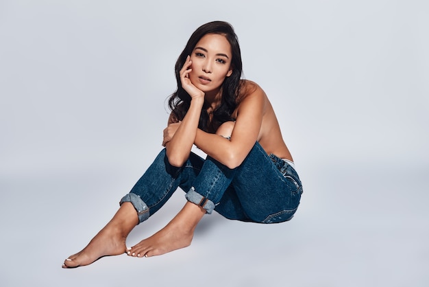 Profiter de sa beauté. Jolie jeune femme regardant la caméra en position assise sur fond gris