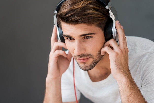 Profiter de sa musique préférée. Beau jeune homme au casque écoutant de la musique assis sur fond gris