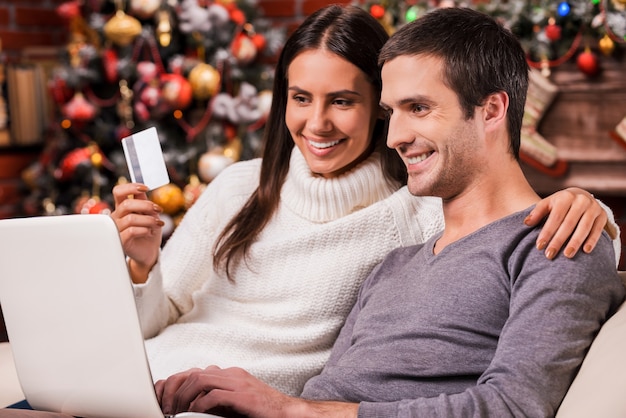 Profiter des soldes de Noël. Beau jeune couple achetant en ligne tout en utilisant un ordinateur avec un arbre de Noël en arrière-plan