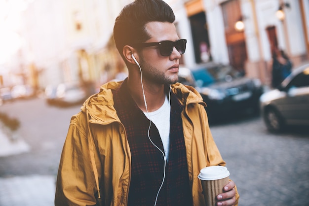 Profiter de la vie en ville. Beau jeune homme au casque portant une tasse de café en marchant le long de la rue de la ville