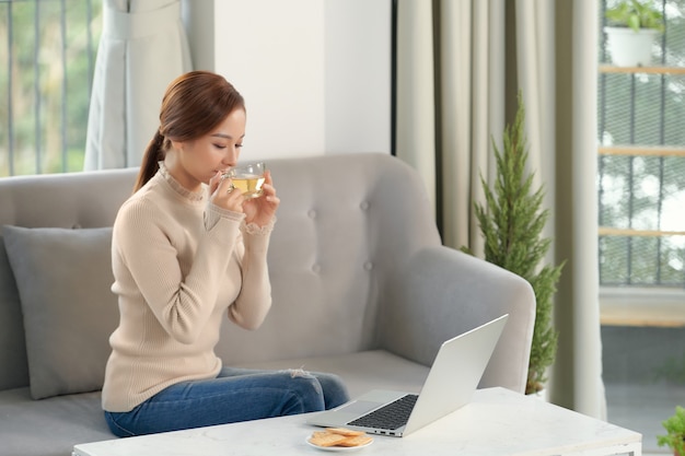 Profitez du week-end. Fille asiatique se relaxant, buvant du café et utilisant un ordinateur portable, assise sur un canapé à la maison