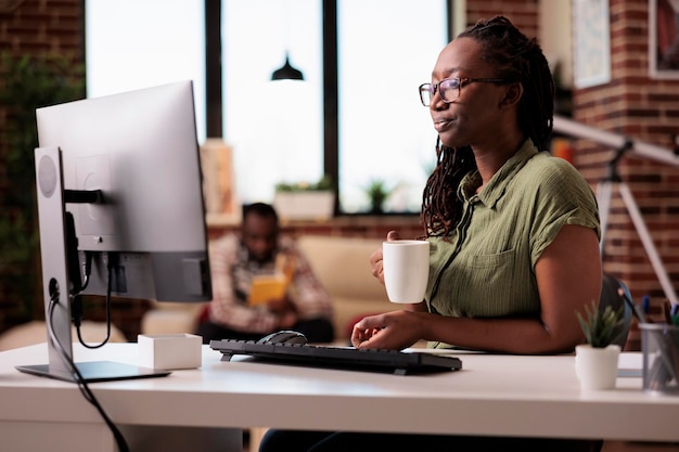 Programmateur à la maison se détendant à l'aide d'un ordinateur pour diffuser des séries en ligne tout en buvant du café ou du thé dans le salon. Freelancer afro-américain passant son temps libre à regarder du contenu vidéo sur l'écran de PC.