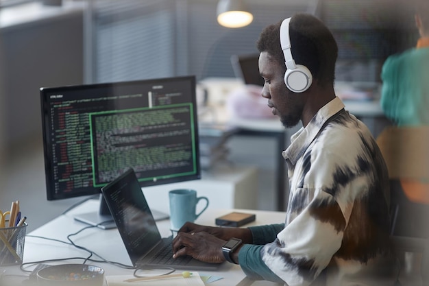 Programmeur afro-américain dans des écouteurs tapant sur un ordinateur portable sur son lieu de travail