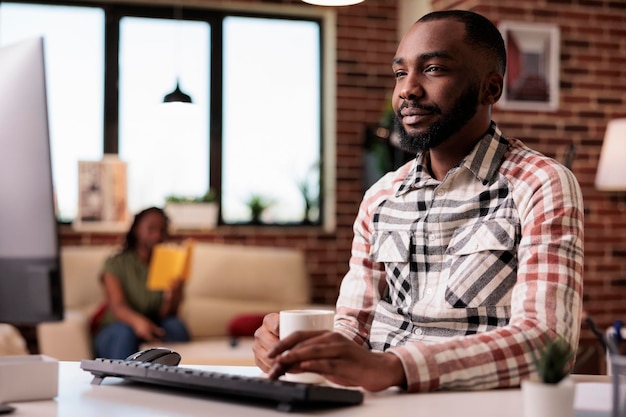 Programmeur afro-américain souriant se relaxant en tenant une tasse de café et en parcourant les médias sociaux sur ordinateur avec sa petite amie lisant sur un canapé. Créateur de contenu travaillant à domicile en faisant une pause au bureau.