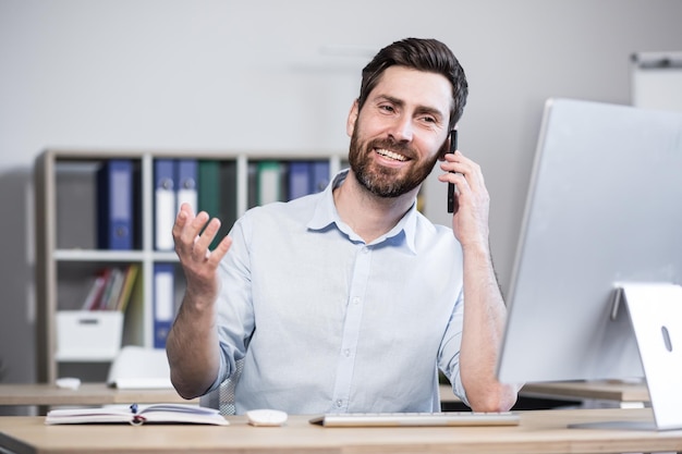 Programmeur masculin réussi travaillant dans le bureau à l'ordinateur parlant à des collègues au téléphone en souriant