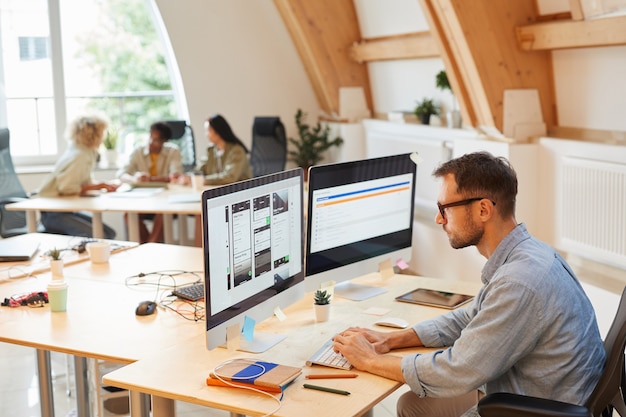 Programmeur sérieux assis sur son lieu de travail à l'aide d'ordinateurs dans son travail, il développe de nouveaux logiciels au bureau avec ses collègues