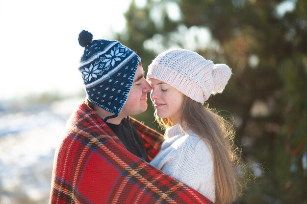 Promenade hivernale à travers les bois. Le gars avec la fille embrassée enveloppé dans un plaid à carreaux rouge