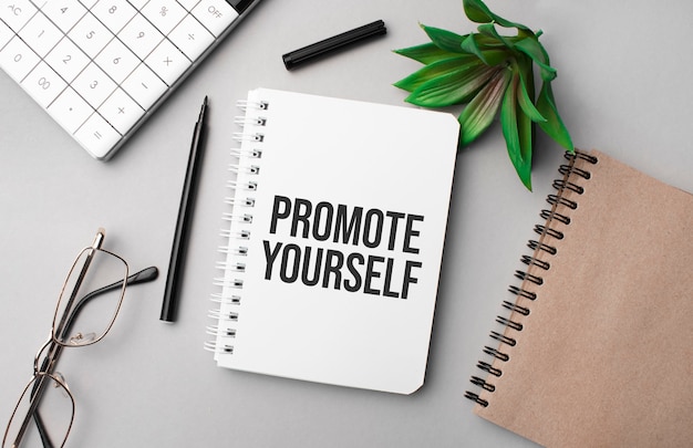 PROMOTE YOURSELF est écrit dans un cahier blanc avec calculatrice, bloc-notes de couleur artisanale, plante, marqueur noir et lunettes.