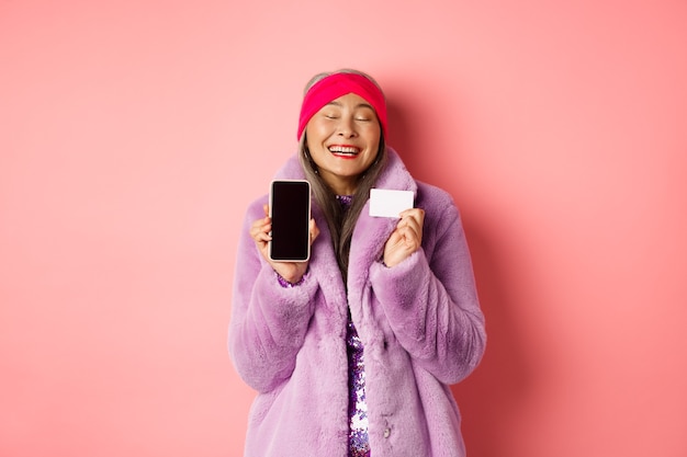 Promotion spéciale. Femme âgée asiatique excitée montrant un écran de smartphone vierge et une carte de crédit en plastique, vérifiant l'offre en ligne, fond rose