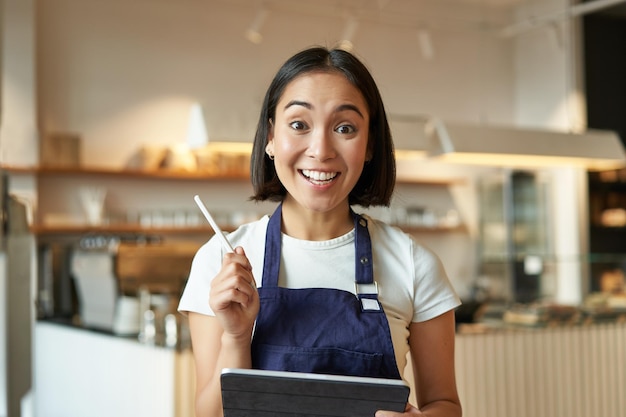 Propriétaire de café entrepreneure asiatique enthousiaste avec tablette portant l'uniforme d'atelier debout avec onglet