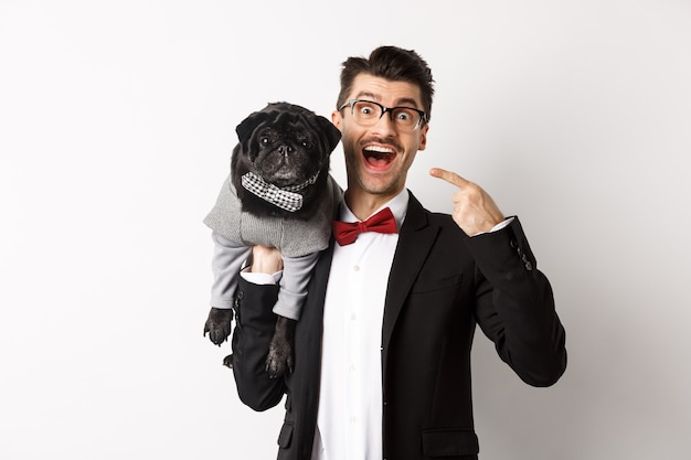 Propriétaire de chien étonné montrant son mignon carlin noir, souriant heureux, chiot en costume, blanc.