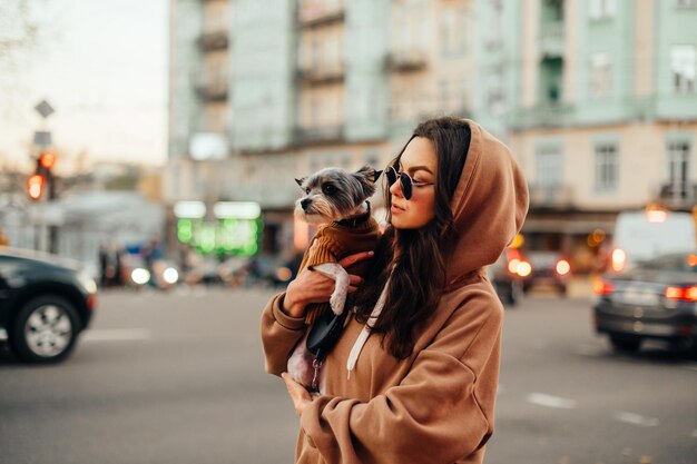 une propriétaire de chien en tenue décontractée et dans des tons se tient dans une rue animée de la ville avec de la circulation