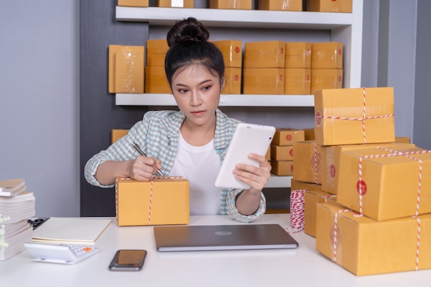 Propriétaire d&#39;une petite entreprise en ligne, femme travaillant avec une tablette numérique, prépare des boîtes à colis pour livraison