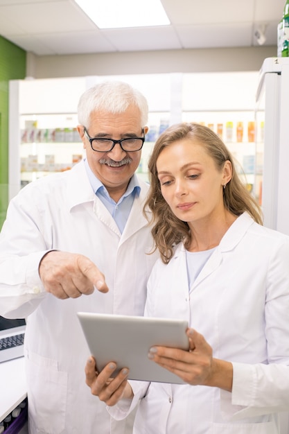 Propriétaire de pharmacie senior pointant sur l'écran du pavé tactile tout en montrant son jeune assistant nouveau médicament dans leur base informatique