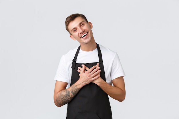 Propriétaires de petites entreprises, café et concept de personnel. Amical, heureux et ravi, beau barista, le serveur en tablier noir apprécie le geste mignon, incline la tête et tient la main sur le cœur heureux