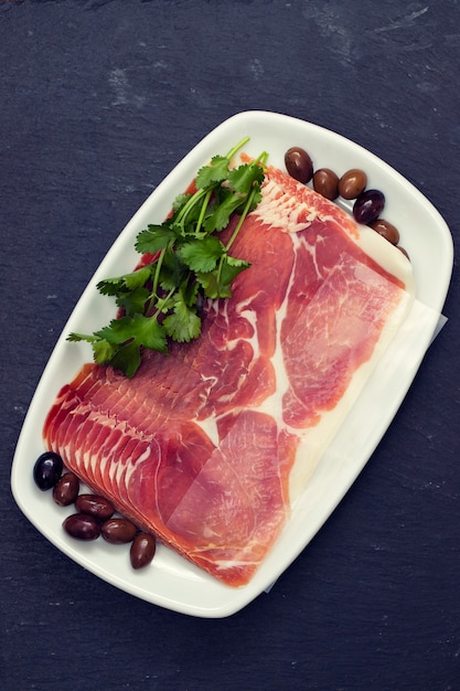 Prosciutto aux olives et persil sur plat blanc sur surface en céramique noire