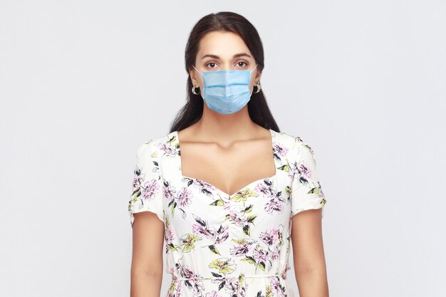 Protection contre les maladies contagieuses, coronavirus. Portrait d'une jeune femme calme avec un masque médical chirurgical en robe blanche debout et regardant la caméra. studio intérieur tourné isolé, fond gris.