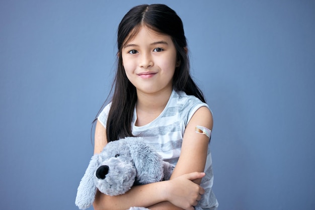 Protéger nos enfants Photo d'une adorable petite fille assise dans le studio après avoir reçu son vaccin Covid et tenant un ours en peluche