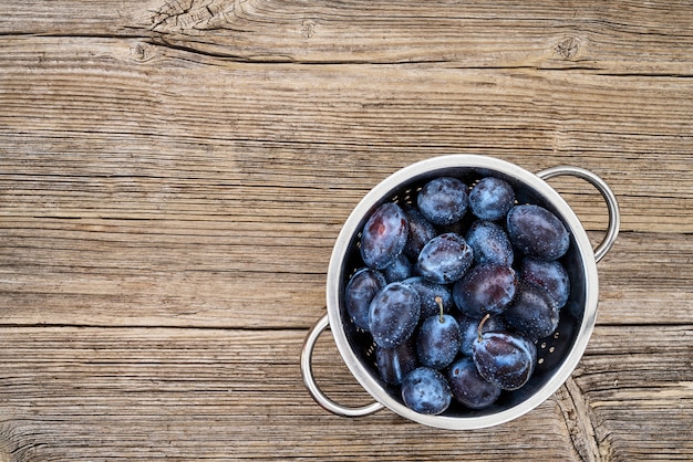 Prunes bleues fraîches dans une passoire rustique sur une vieille table en bois. Alimentation biologique. Vue de dessus, espace de copie.