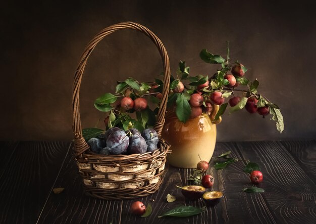 Prunes dans un panier et branches de pomme dans un vase Nature morte aux prunes et pommes