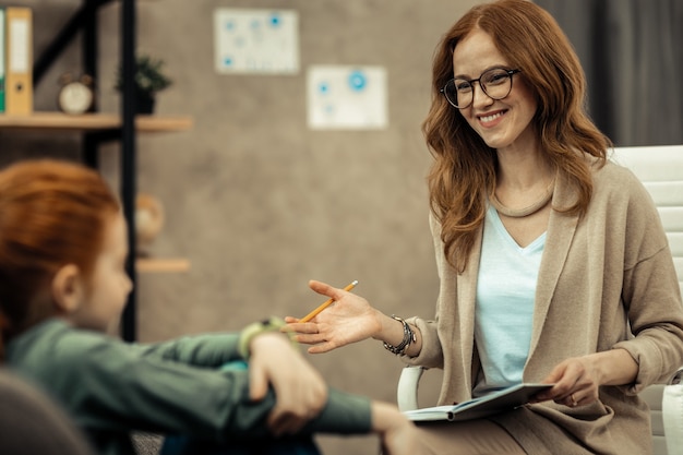 Psychologue expérimenté. Jeune femme intelligente regardant ses notes tout en ayant une session avec un enfant