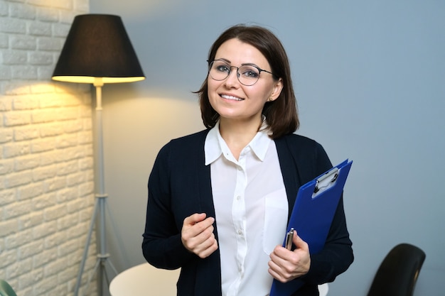 Psychologue de femme mûre, psychiatre, travailleur social avec le presse-papiers regardant la caméra, intérieur de bureau de fond