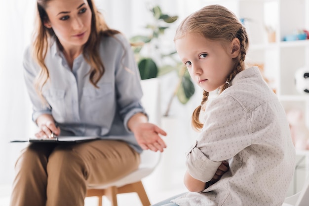 Psychologue parlant à un enfant en colère avec les bras croisés pendant qu'elle regarde la caméra