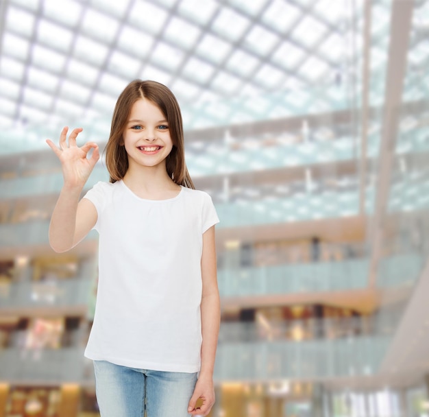publicité, enfance, geste, consommation et personnes - petite fille souriante en t-shirt blanc vierge montrant le signe ok sur fond de centre commercial