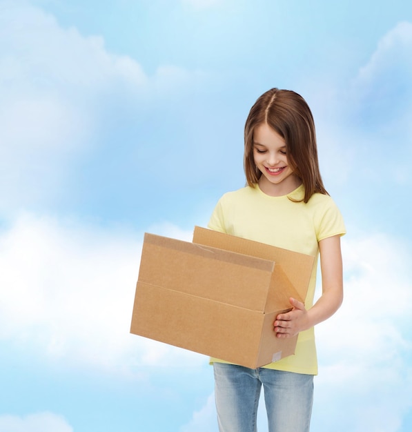 publicité, enfance, livraison, courrier et personnes - petite fille souriante tenant une boîte en carton ouverte et la regardant sur fond nuageux