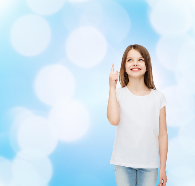 publicité, geste, enfance et personnes - petite fille souriante en t-shirt blanc vierge pointant le doigt vers le haut sur fond bleu