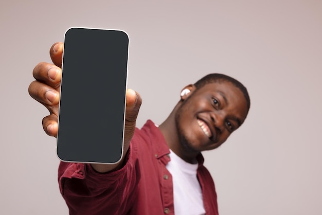 Publicité mobile Homme noir ravi pointant vers un téléphone portable avec un écran blanc vide sur une maquette de fond de studio bleu pour une application ou un site Web Modèle d'affichage de téléphone portable