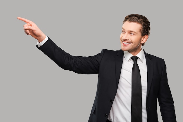 Publicité de votre produit. Jeune homme confiant en tenue de soirée regardant loin et souriant tout en pointant du doigt et debout sur fond gris