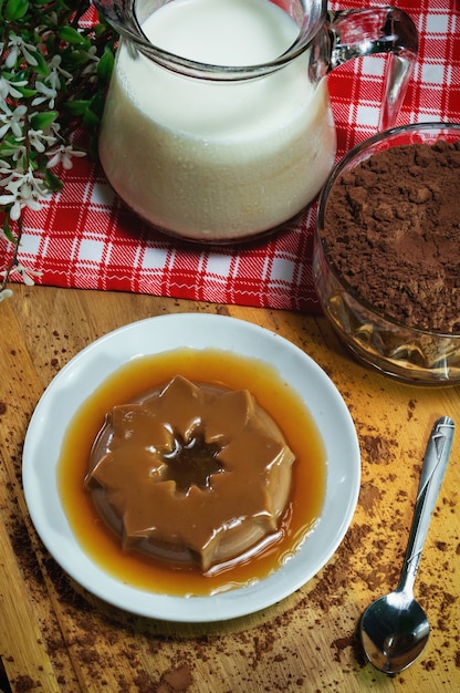 Pudding au chocolat sur assiette en porcelaine. Pot de lait et pot en verre avec du chocolat en po. Base en bois avec tissu en damier. Fond noir.