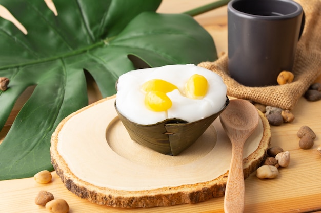 Pudding thaï à la noix de coco et aux graines de Ginkgo biloba, Desserts enveloppés dans des feuilles de bananier. Les Thaïlandais appellent "Khanom Tako". Tako avec du Ginkgo placé sur une soucoupe en bois et une cuillère en bois, avec du thé dans un verre noir.