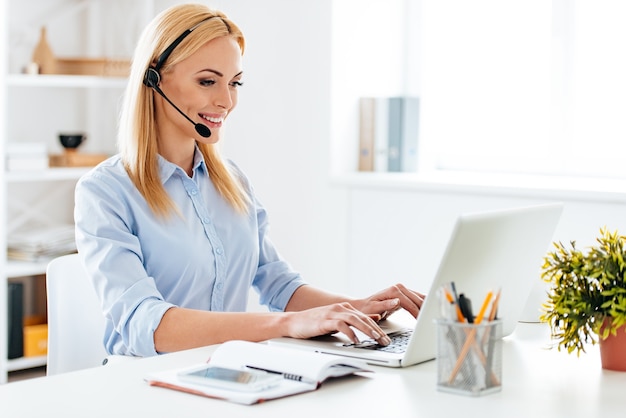 Puis-je vous aider? Joyeuse jeune femme belle dans des écouteurs travaillant sur un ordinateur portable et souriante assise sur son lieu de travail