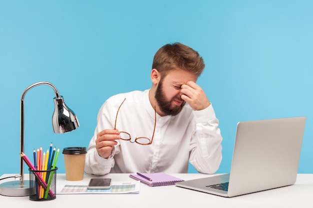 Épuisé homme stressé employé de bureau frottant fatigué des yeux de travail d'ordinateur portable repoussant les lunettes assis sur le lieu de travail problèmes de surmenage avec la vision prise de vue en studio intérieur isolé sur fond bleu