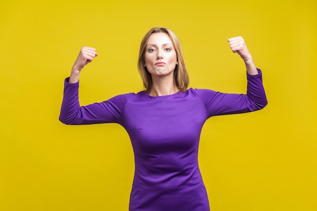 Puissance et force intérieure féminine. Portrait d'une femme d'affaires fière et prospère vêtue d'une élégante robe violette démontrant des biceps, se sentant indépendante et confiante, le féminisme. tourné en studio isolé sur jaune