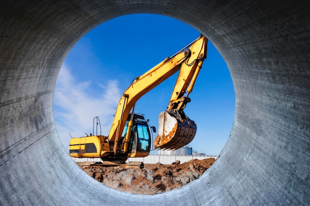 Une puissante excavatrice à chenilles creuse le sol contre le ciel bleu Travaux de terrassement avec de l'équipement lourd sur le chantier de construction Vue à travers un grand tuyau en béton