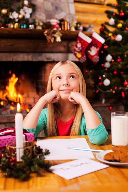 Qu'est-ce que je souhaite ? Jolie petite fille rêvant d'une journée assise à la maison avec un arbre de Noël et une cheminée en arrière-plan