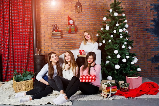 Quatre amies mignonnes portent des pulls chauds, des pantalons noirs et des chapeaux de père Noël contre un arbre du nouvel an avec une décoration de Noël au studio.