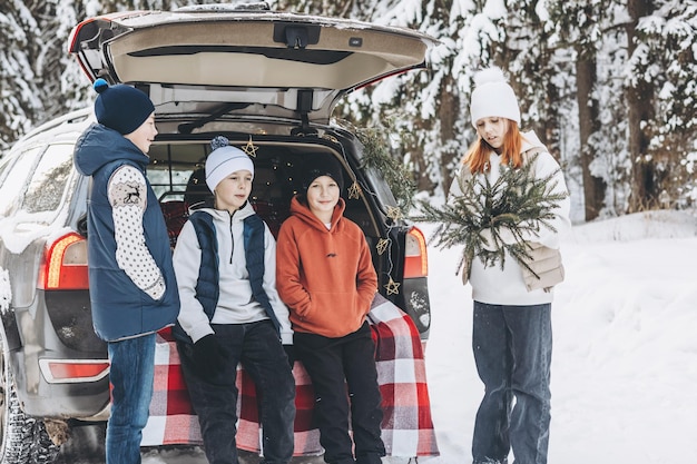 Quatre amis adolescents garçons et filles avec thermos assis dans le coffre d'une voiture décorée pour Noël et le Nouvel An Pique-nique d'hiver dans la forêt enneigée Voyage en voiture et voyages locaux Amis traînant ensemble